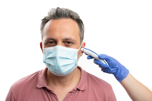 Man wearing surgical mask having temperature measured in ear with laser thermometer by person wearing medical latex gloves as covid19 symptom concept isolated on white background